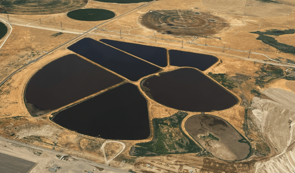 An aerial view of 6 large dark pools of manure sit on yellow earth near roads that look as small as a length of yarn.