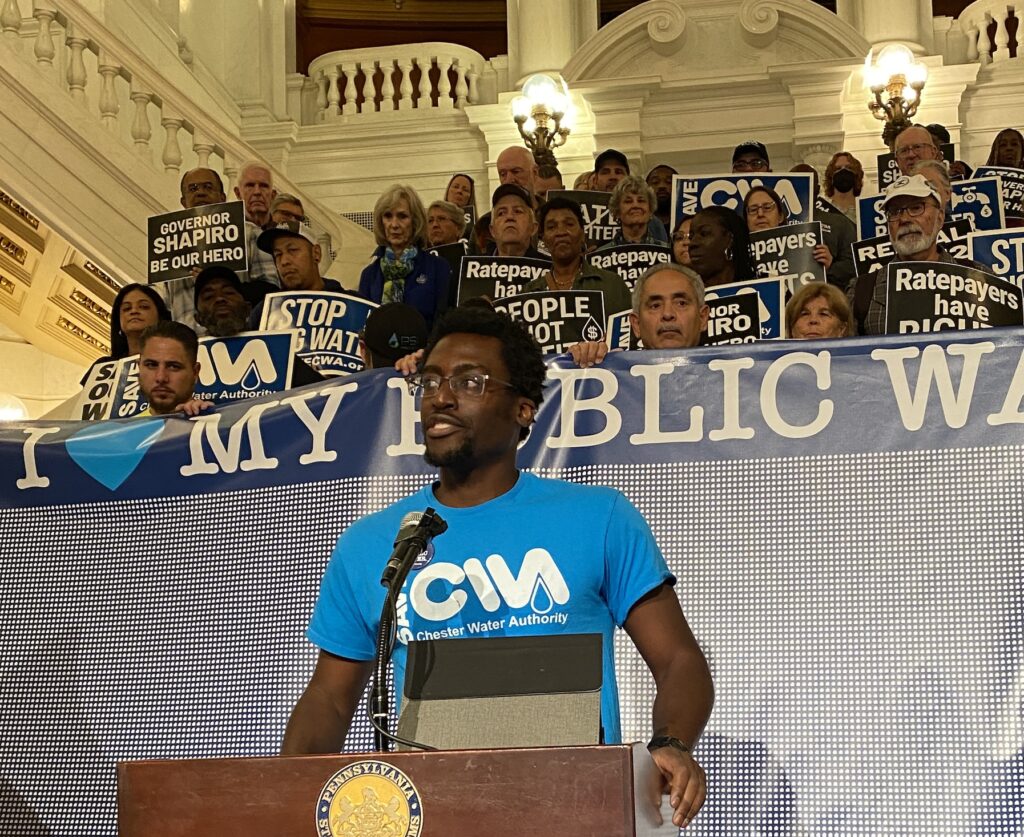 Inside the white halls of the Capitol building, Kofi stands in front of a podium, with dozens of advocates standing behind him on steps. The front row of advocates holds a banner reading 'I heart my public water."