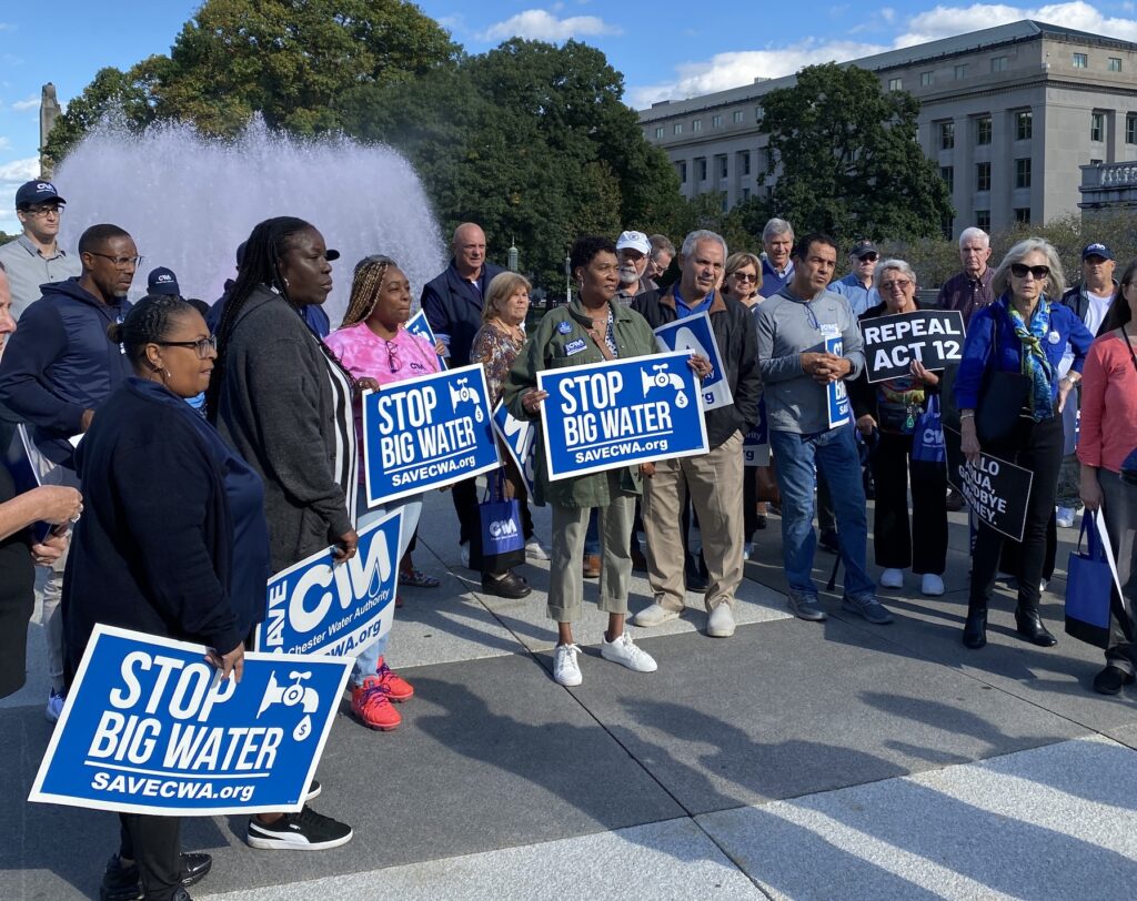 Dozens of people stand in front of a fountain outside the Capitol holding blue signs that read "Stop Big Water," "Repeal Act 12."