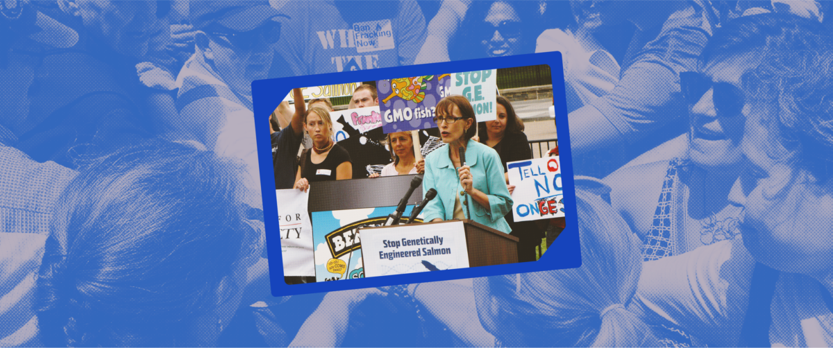 Wenonah speaks behind a podium surrounded by protestors holding signs.