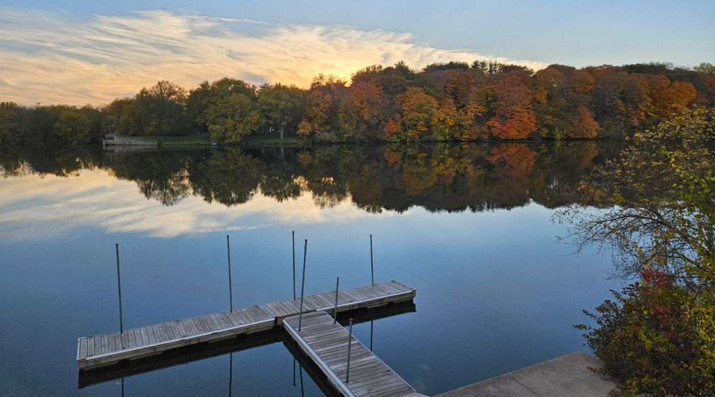 A lack in Backbone State Park, with a jetty jutting out from the bottom of the photo and a line of orange and green trees on the lake's far shore.