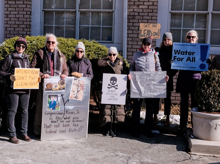 Seven protestors in winter coats stand in front of a brick building and green hedges. They hold signs reading "Get the Lead Out!" "Hey Rep. Kean, don't fight clean water!" and "Water for All!"