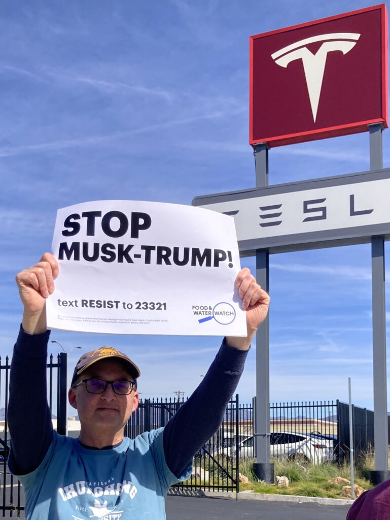 Mark holds a sign that reads "Stop Musk-Trump!" in front of a sign with the Tesla logo.